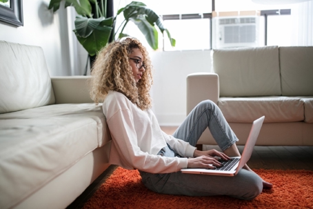 A person with blonde curly hair sits on an orange rug and works on a laptop