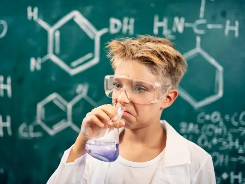 A young boy wears safety goggles and a lab coat, grimacing as he smells a purple liquid
